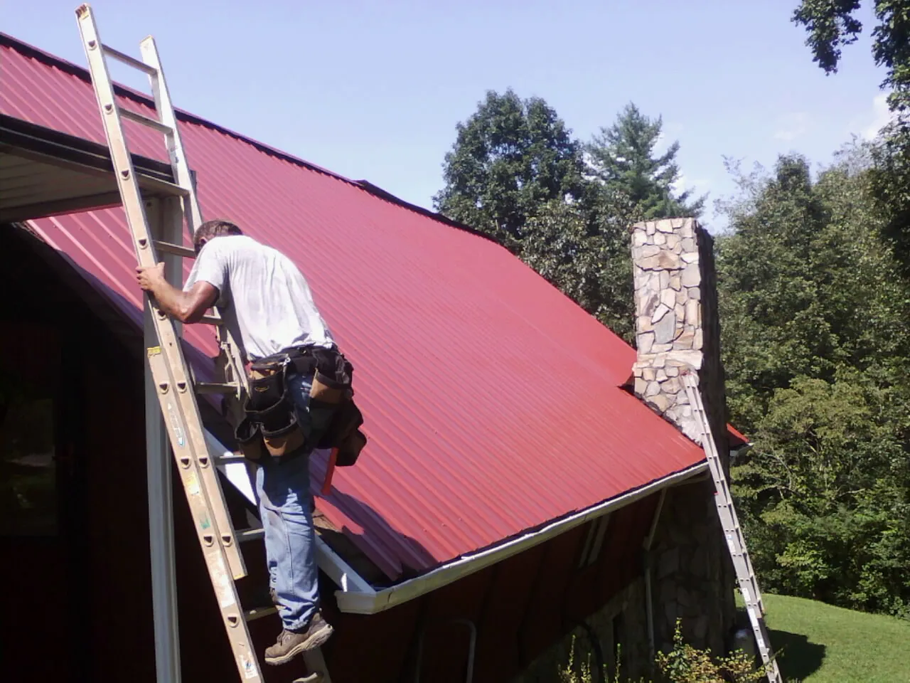 Roofer on a ladder beside a red metal roof and stone chimney — Soffit & Fascia Repair by Roofing Brothers in Buchanan Dam